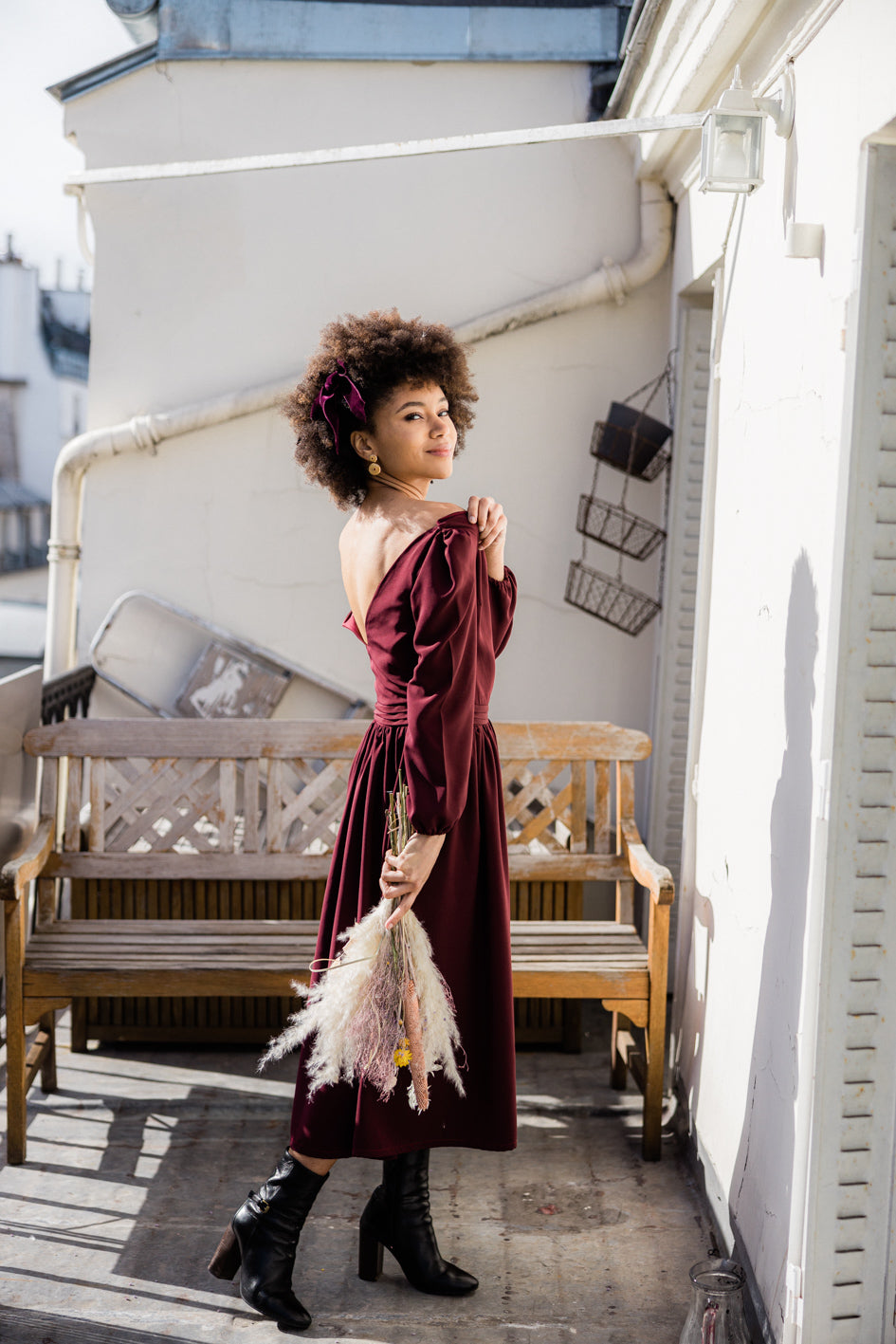 Robe bordeaux dos nu en crêpe de soie fluide, manches longues bouffantes, confection sur mesure dans l’atelier de couture parisien. Création élégante et féminine réalisée à la commande, mise en valeur en extérieur sous la lumière naturelle.