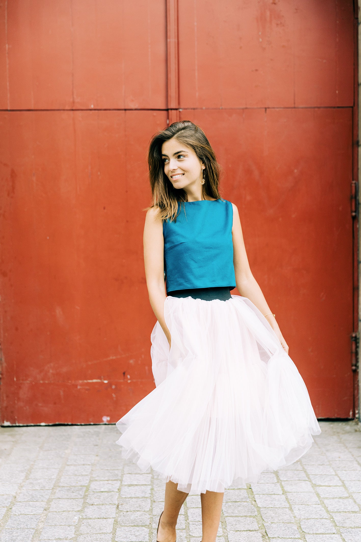 Mannequin qui porte l'ensemble Carrie. Un tutu en tulle de soie rose pastel marqué par une ceinture large et noire ; un crop top bleu canard col rond ajusté à la poitrine. Ensemble porté en pleine lumière naturelle. 